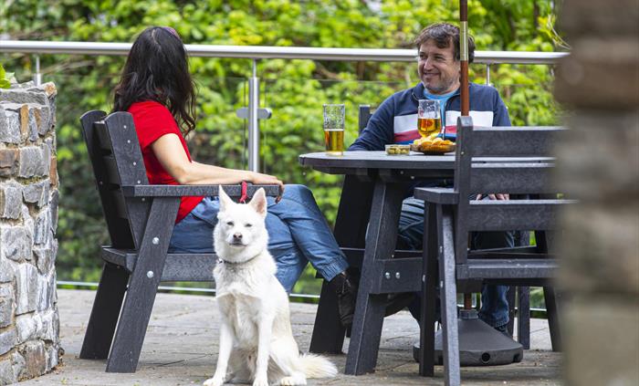 A white dog sits in front of a couple enjoying drinks at a black patio table with stone walls and greenery in the background